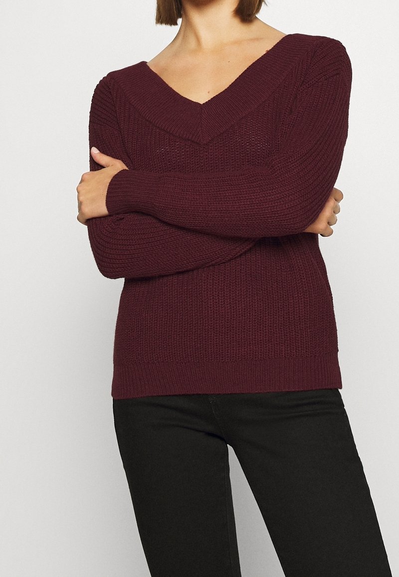 Personne portant un pull en maille col en V couleur bordeaux, les bras croisés, associé à un pantalon noir, sur un fond clair uni.