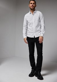 Bílá košile na knoflíky s jemným černým potiskem, v kombinaci s černými úzkými kalhotami a robustními černými botami. Jednoduchý, moderní design.