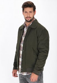 Oliven grøn jakke med krave, struktureret stof og frontlukning med knapper. Båret over en rutete skjorte, viser grå jeans. Ankerlogo detalje.