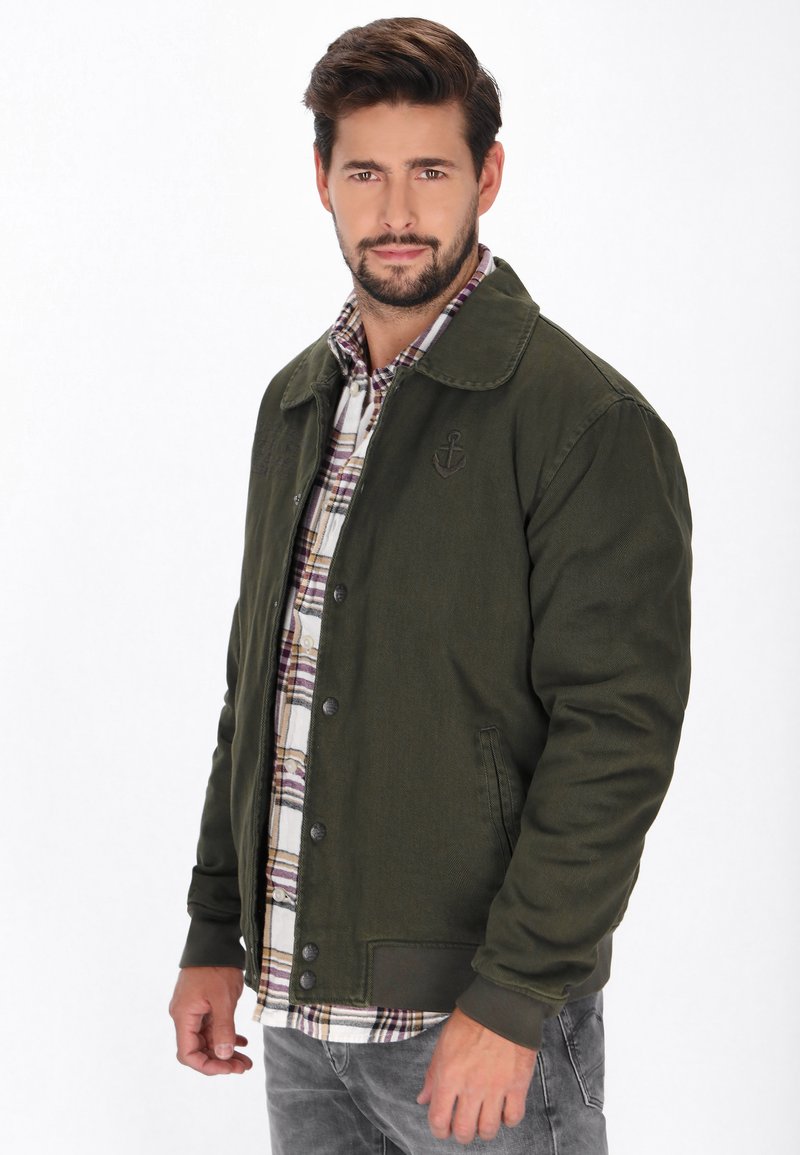 Oliven grøn jakke med krave, struktureret stof og frontlukning med knapper. Båret over en rutete skjorte, viser grå jeans. Ankerlogo detalje.