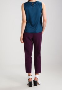 Zielona bezrękawnik z detalem w kształcie dziurki na plecach, w połączeniu z fioletowymi, zwężającymi się spodniami i białymi sandałami na obcasie, umiejscowiony na neutralnym tle.