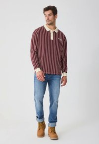 Ilgas rankovės polo marškinėliai bordo spalvos su baltomis vertikaliomis juostomis, su šviesios kremine apykakle ir rankogaliais, derinami su mėlynais džinsais ir rusvais batais.