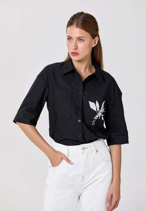 Femme aux cheveux bruns portant une chemise noire à boutons avec un motif floral blanc sur la poche et un pantalon blanc taille haute, debout avec une main dans la poche.