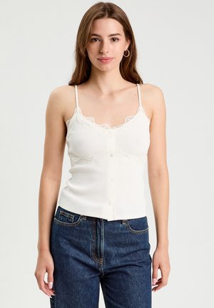 Camisole bianca a coste con rifinitura in pizzo e dettagli con bottoni sulla parte anteriore, abbinata a jeans blu scuro a vita alta su una modella.