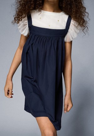 Niña con un vestido tipo pichi azul marino sobre una camiseta blanca con mangas transparentes con volantes y pequeños adornos en forma de puntos dorados.