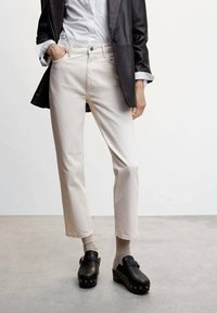 Jean beige clair avec une coupe droite, comportant des poches avant et un ourlet brut, associés à des sabots noirs et des chaussettes crème.