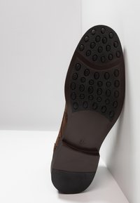 Semelle d'une chaussure habillée marron avec des patins en caoutchouc noir et des clous ovales, taille 41, positionnée contre un coin blanc.