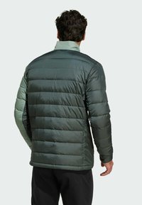 Giacca a vento verde scuro con design trapuntato, polsini elasticizzati e colletto in un verde chiaro a contrasto. Il materiale sembra liscio e isolante.