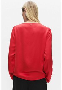 Punainen satiinipaita, jossa on sileä pinta, pyöreä kaula-aukko, pitkät hihat ja nappikoristelu hiharesoreissa; takana on pieni lenkki.