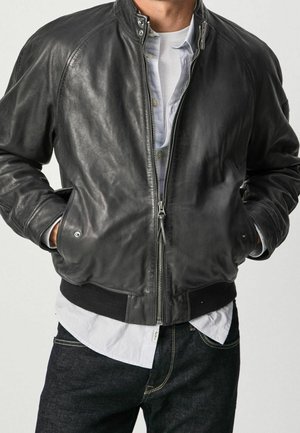 Mann trägt schwarze Lederbomberjacke über weißem Hemd und dunklen Jeans, Hände in den Taschen, Jacke teilweise geöffnet, steht vor einfachem Hintergrund.