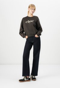 Mujer joven con cabello rubio en una coleta, vistiendo un sudadera negra con el texto "choc", jeans oscuros de pierna ancha y zapatillas negras, de pie con las manos en los bolsillos.