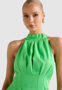 Γυναίκα με ξανθά μαλλιά σε κομψό κότσο, φορώντας χρυσούς κρίκους σκουλαρίκια και φωτεινό πράσινο πλισέ φόρεμα με λαιμόκοψη halter, πάνω σε λευκό φόντο.