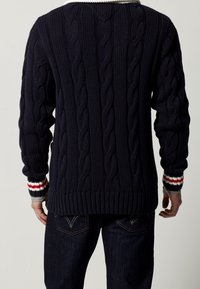 Marinblå stickad tröja med ett kabelmönster, ribbade ärmslut med röda och vita accenter, samt en avslappnad passform, sedd från baksidan.