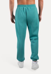 Turquoise sweatpants met een elastische tailleband en taps toelopende boorden. De stof is zacht en glad. Draagt met witte sneakers.