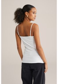 Sieviete ar cirtainu asti, vilkdama baltu topu ar plānām siksnām un melnām džinsēm, stāv profila pozā uz neitrāla fona.