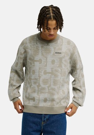 Jonge man met gevlochten haar, die een beige trui met patroon en blauwe spijkerbroek draagt, kijkt naar zijn trui.