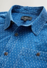 Cămașă cu nasturi din denim albastru, cu dungi verticale albe, prevăzută cu un buzunar pe piept și o guler standard. Detaliu de închidere cu nasturi negri.