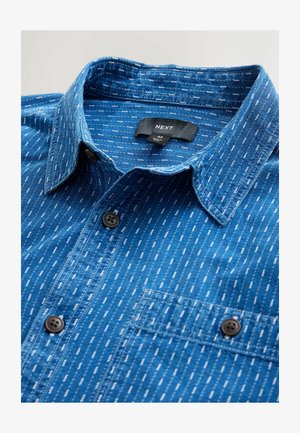 Gombos ing kék farmert textilel, fehér függőleges csíkokkal, mellzsebbel és normál gallérral. Fekete gombzár részlet.