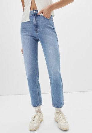 Personne portant un jean droit taille haute bleu clair, des baskets blanc cassé et un top court blanc, une main dans la poche.