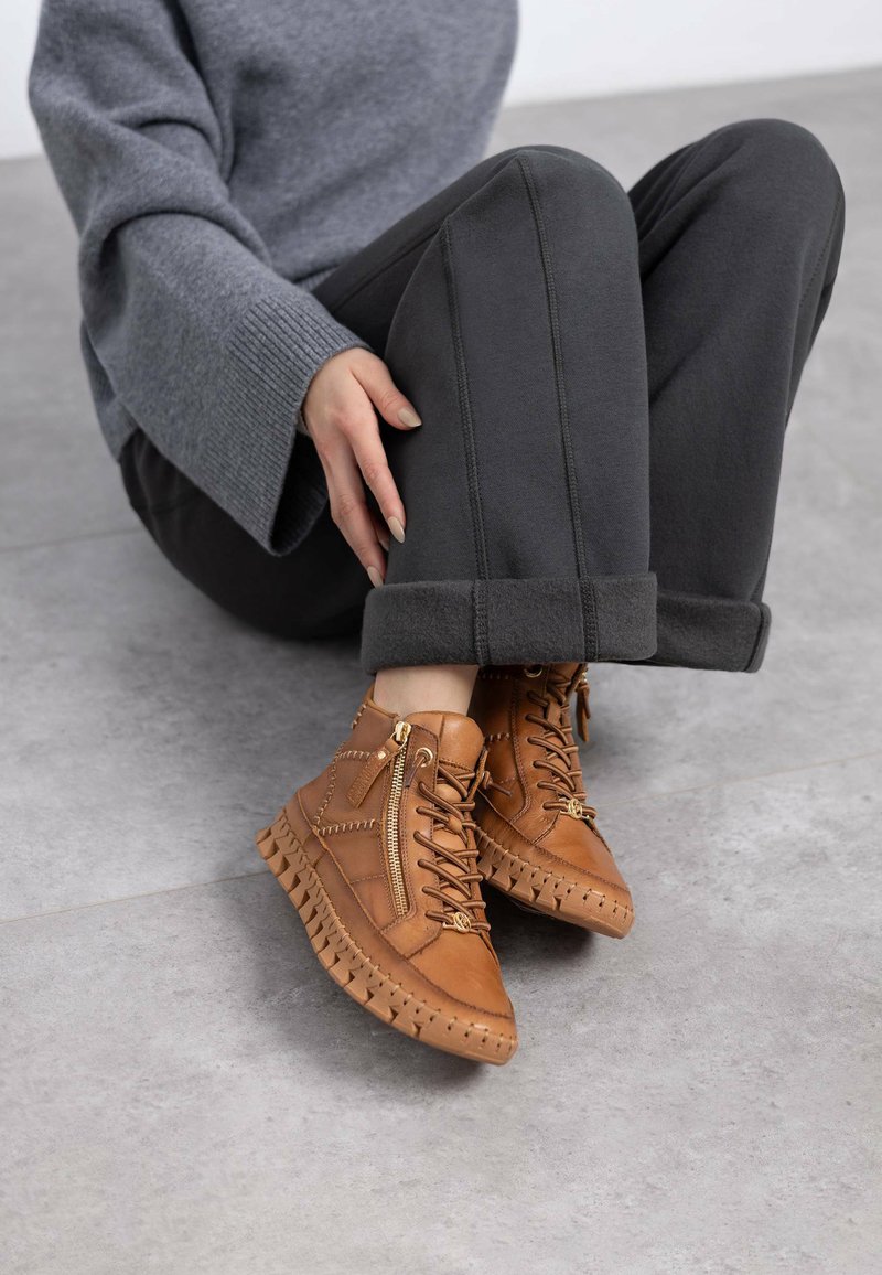 Botas de tobillo de cuero marrón con cremalleras laterales, suelas texturizadas y detalles de costura; combinadas con pantalones grises y un suéter gris.