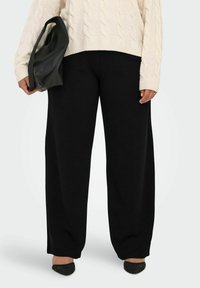 Person, die eine schwarze weite Hose, einen cremefarbenen Kabelstrick-Pullover, schwarze geflochtene spitze Schuhe trägt und eine schwarze Leder-Clutch hält.