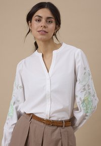 Femme portant un chemisier blanc boutonné avec broderie florale sur les manches, un pantalon plissé beige et une ceinture tressée marron sur fond uni.