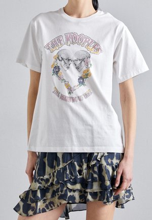 Camiseta de algodón blanco con un diseño gráfico de dos calaveras rodeadas de flores. Combinada con una falda con volantes y estampado de leopardo en azul y verde.