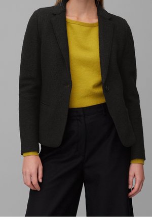 Person iført en tætsiddende sort blazer over en sennepsgul sweater og løsthængende sorte bukser mod en ensfarvet grå baggrund.