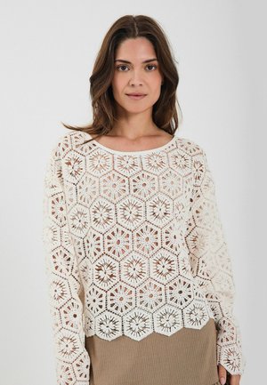 Koronkowy sweter w kolorze kremowym z heksagonalnymi wzorami, z otwartym splotem i falbaniastym dołem. Długie rękawy i szeroki dekolt.