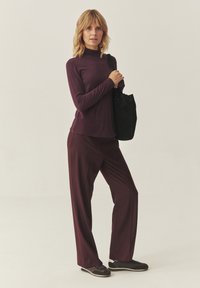 Burgundi hosszú ujjú felső és hozzáillő, bővülő nadrág, sima anyagból készült. A modellt egy fekete táska és sötét sportcipő öltözteti.