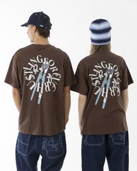 Braune Baumwoll-T-Shirts mit einem Grafikdruck, der eine Schere auf dem Rücken zeigt. Kombiniert mit blauen Jeans. Flachkappen und eine Mütze werden getragen.