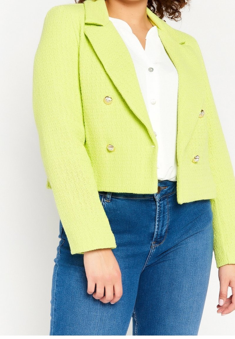 Žena v krátké texturované blůze limetkově zelené barvy se zlatými knoflíky, bílé košili a modrých džínách, stojící na bílém pozadí.