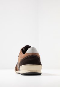 Baskets en daim marron avec accents en cuir, coiffe de talon grise, semelle blanche et ligne de ventilation latérale. Surface texturée avec détails de couture.