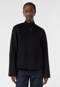Frau mit geflochtenem Haar trägt eine schwarze Zip-Jacke mit großen Fronttaschen und dunklen Hosen und steht vor einem schlichten weißen Hintergrund.