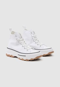 Zapatillas altas de lona blanca con una suela de goma gruesa y texturizada y detalles en negro, con ojales de metal y cordones en la parte delantera.