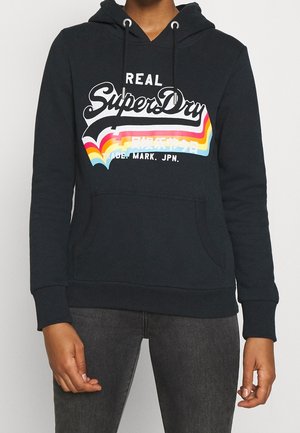 Czarna bluza z kapturem z przednią kieszenią, na której znajduje się napis "Real SuperDry" oraz wielokolorowy paskowany wzór, noszona w zestawie z ciemnoszarymi dżinsami.