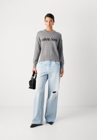Hallhalli hallituspüksi, millel on "calvin klein" logo, koos heledate siniste laiade pükstega, millel on kulumise jäljed. Mudel hoiab musta kotti.