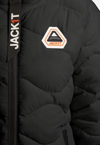 Giacca a vento nera con texture trapuntata, che presenta un evidente logo esagonale cucito e una cerniera laterale con una linguetta brandizzata.