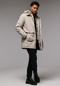 Parka acolchada beige con capucha, que presenta un cierre con cremallera y botones a presión en el frente, bolsillos laterales y un corte relajado, complementado con pantalones y botas negras.