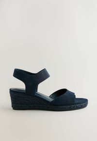 Mørkeblå kilehæle i ruskind med åben tå og bred elastisk ankelrem. Tekstureret vævet hæl tilføjer højde og stabilitet.