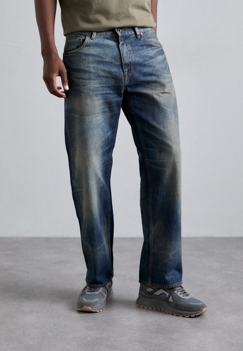 Jean en denim bleu délavé avec une coupe straight-leg, présentant un design classique à cinq poches et des coutures contrastantes. Portés avec des baskets grises.