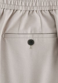 Tissu gris clair avec une ceinture élastique, une poche arrière unique avec un bouton noir, et des motifs texturés subtils.