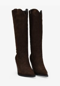 Botas altas de ante marrón hasta la rodilla con puntera afilada, borde superior con forma de ondas y cremallera lateral. Con tacón de cuña y suela plana.