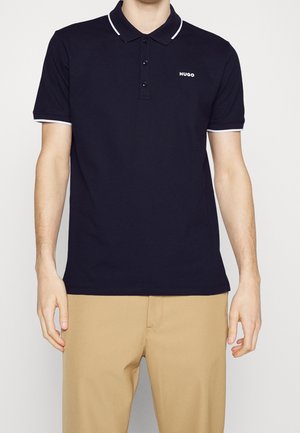 Homme portant un polo Hugo bleu marine avec une bordure blanche au col et aux manches, associé à un pantalon beige, montré du cou jusqu'aux hanches.