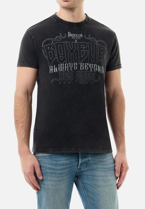 Mann trägt ein ausgewaschenes schwarzes Boxeur des Rues T-Shirt mit dem Schriftzug „Always Beyond Des Rues“ und hellblaue Jeans, steht mit entspannten Armen.