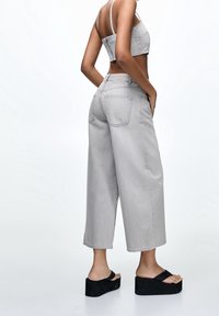 Femme portant un jean gris clair large et court, un haut court gris, et des sandales à plateforme noires, debout les mains dans les poches devant un fond blanc.