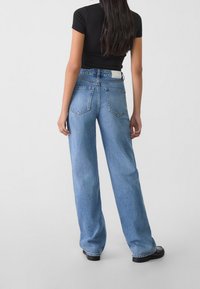 Mujer con cabello largo y oscuro, vistiendo una camiseta negra de mangas cortas, jeans rectos de tiro alto en azul claro y zapatos negros, de pie dando la espalda.