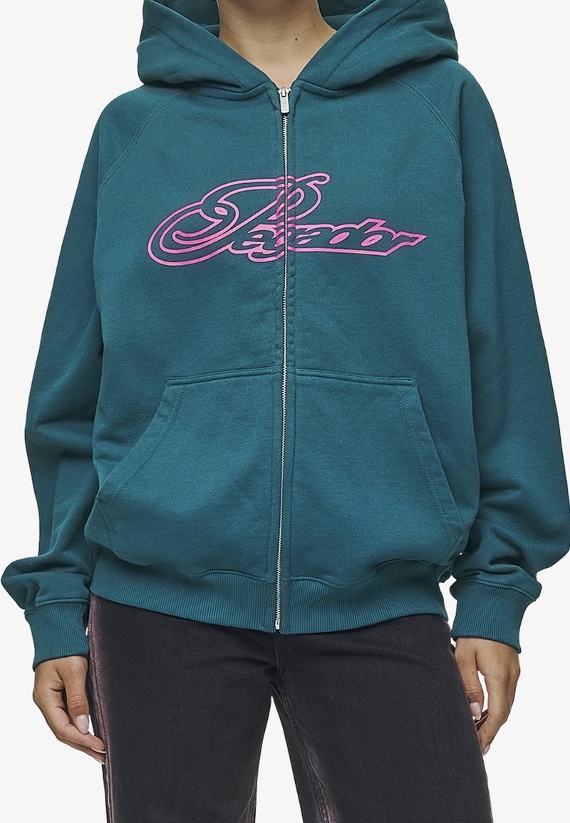 Personne portant un sweat à capuche zippé couleur sarcelle avec un logo "Psyador" rose en écriture cursive et des poches avant, associé à un pantalon noir avec une bande latérale brillante.