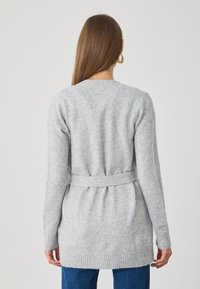 Pilkas megztas kardiganas su šonkačių užlenkimu, elastine juosta ir ilguomis rankovėmis. Minkšta tekstūra ir laisvas siluetas.