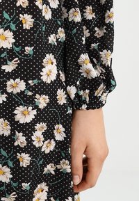 Tissu noir avec des marguerites blanches et des accents verts, agrémenté d'un motif à pois. Détail de manches froncées et texture lisse.